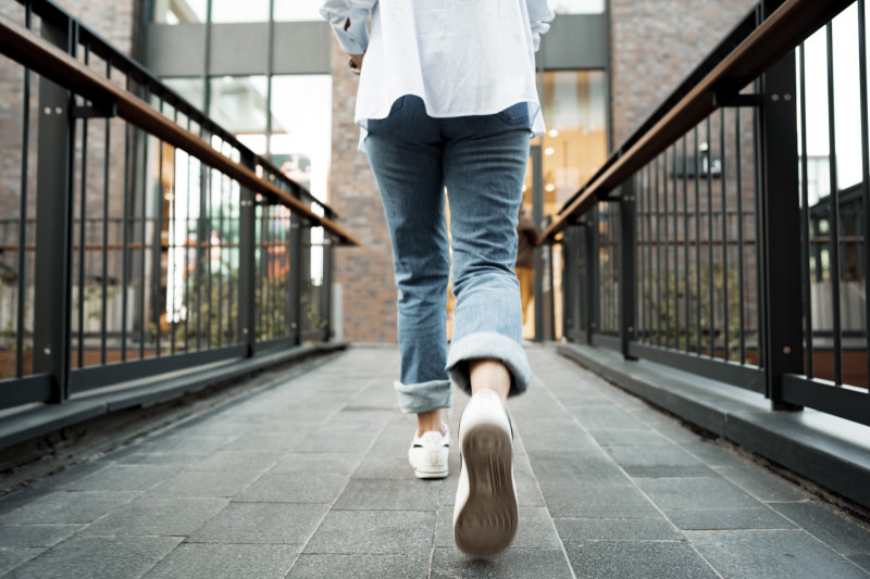 Slim lady in white sneakers, gray pants and white oversized blouse poses against brick wall surroun