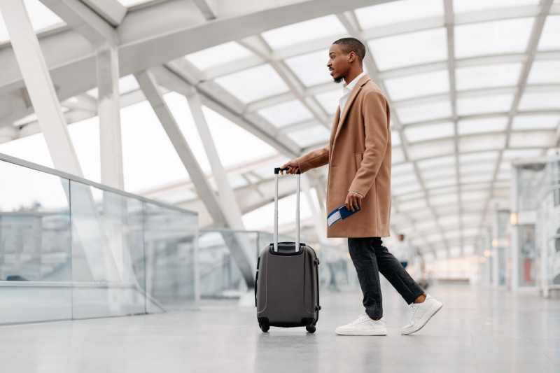 Handsome Black Man Wearing Stylish Coat Walking With Suitcase At Airport